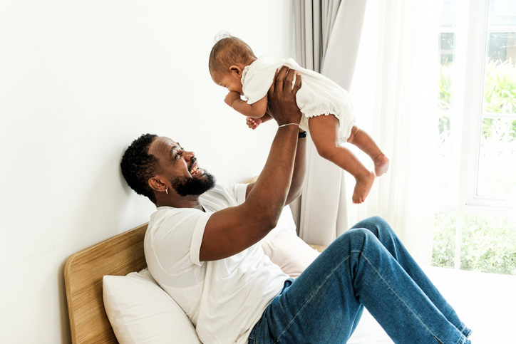 Joyful Black father lifting his baby daughter in a sunlit bedroom, expressing love, bonding, and parenthood. Great for Father's Day, family values, and authentic diverse lifestyle visuals.