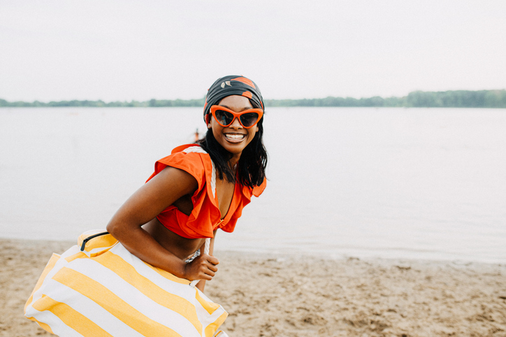 A woman in vibrant orange attire and sunglasses joyfully enjoys a day at the beach, holding a yellow and white striped tote